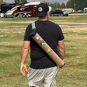 May include: A person wearing a black cap with the number 41 and a black t-shirt, gray shorts, and green and black sneakers. A brown cylindrical carrying case with stickers is slung over the shoulder. The person is walking on a grassy field.