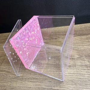 May include: A clear acrylic box with a pink glitter-filled interior panel. The box has a hinged lid with small holes and is angled on a wooden surface. The glitter is a mix of pink, silver, and iridescent shapes.