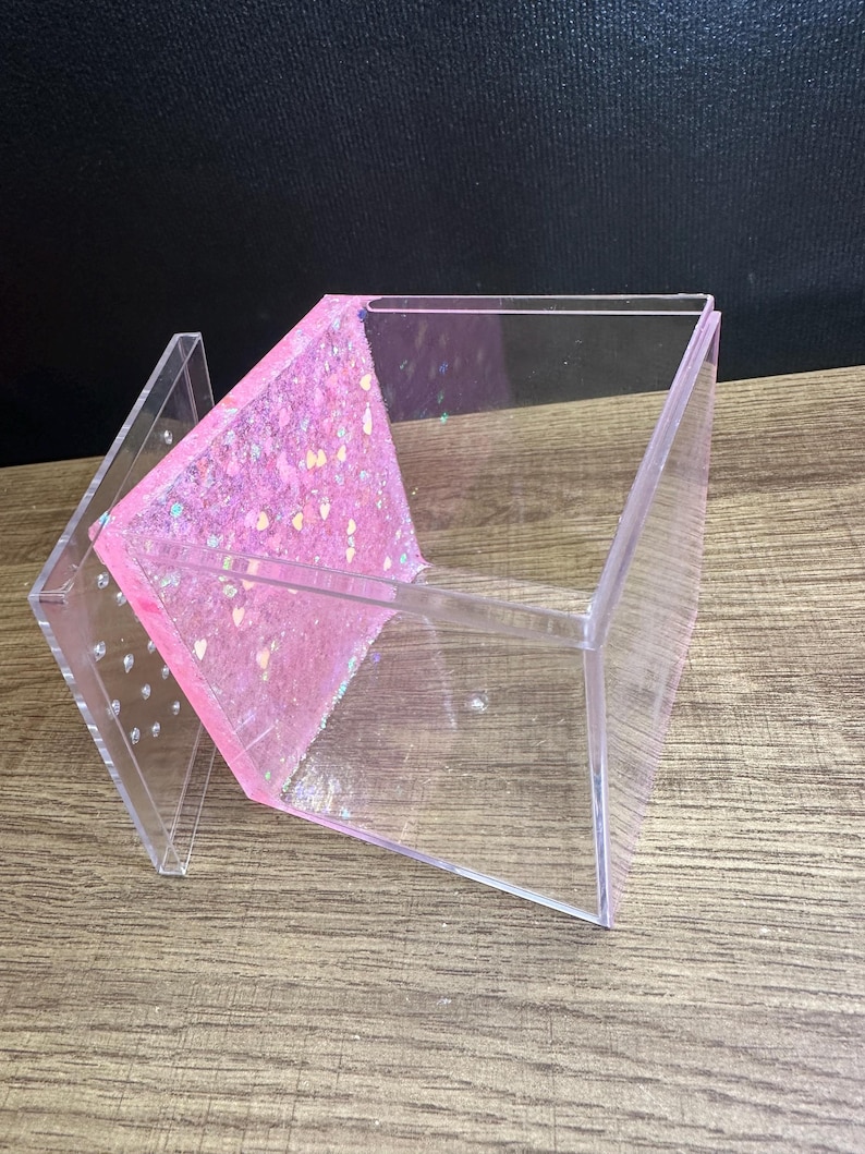 May include: A clear acrylic box with a pink glitter-filled interior panel. The box has a hinged lid with small holes and is angled on a wooden surface. The glitter is a mix of pink, silver, and iridescent shapes.