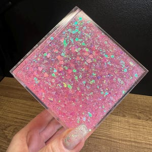 May include: A clear, square container filled with pink glitter and iridescent specks. The glitter is suspended in a clear liquid, creating a shimmering effect. The container is held in a hand.