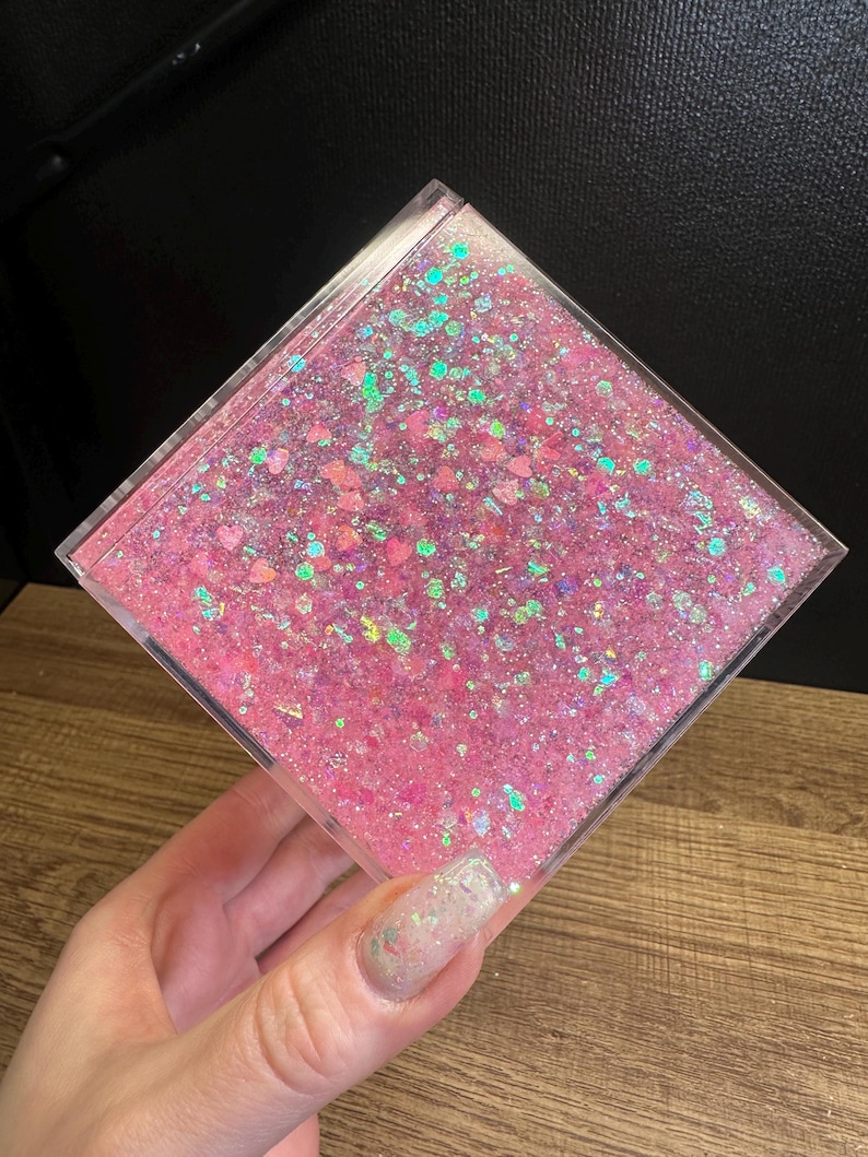 May include: A clear, square container filled with pink glitter and iridescent specks. The glitter is suspended in a clear liquid, creating a shimmering effect. The container is held in a hand.