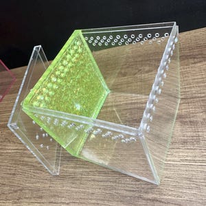 May include: Clear acrylic terrarium with a green interior and small air holes. The terrarium is angled and has a geometric design. The interior has a textured, speckled appearance. The terrarium is designed to house small animals or plants.