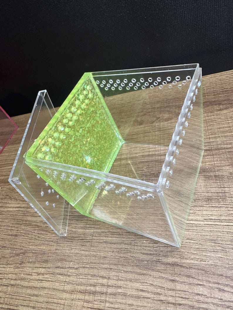 May include: Clear acrylic terrarium with a green interior and small air holes. The terrarium is angled and has a geometric design. The interior has a textured, speckled appearance. The terrarium is designed to house small animals or plants.