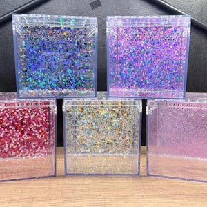 May include: Five clear, square containers filled with colorful glitter. The glitter colors include blue, purple, red, gold, and pink. The containers are made of clear plastic and are arranged in a group.
