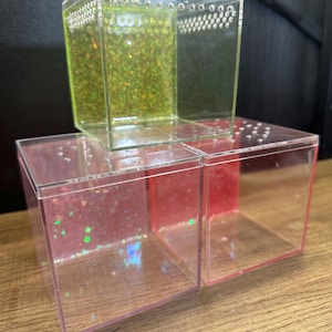 May include: Clear acrylic cube-shaped containers, one stacked on top of two others. The top cube has a green glitter filling, while the bottom cubes have pink and red glitter. Each cube has small holes along the top edge.