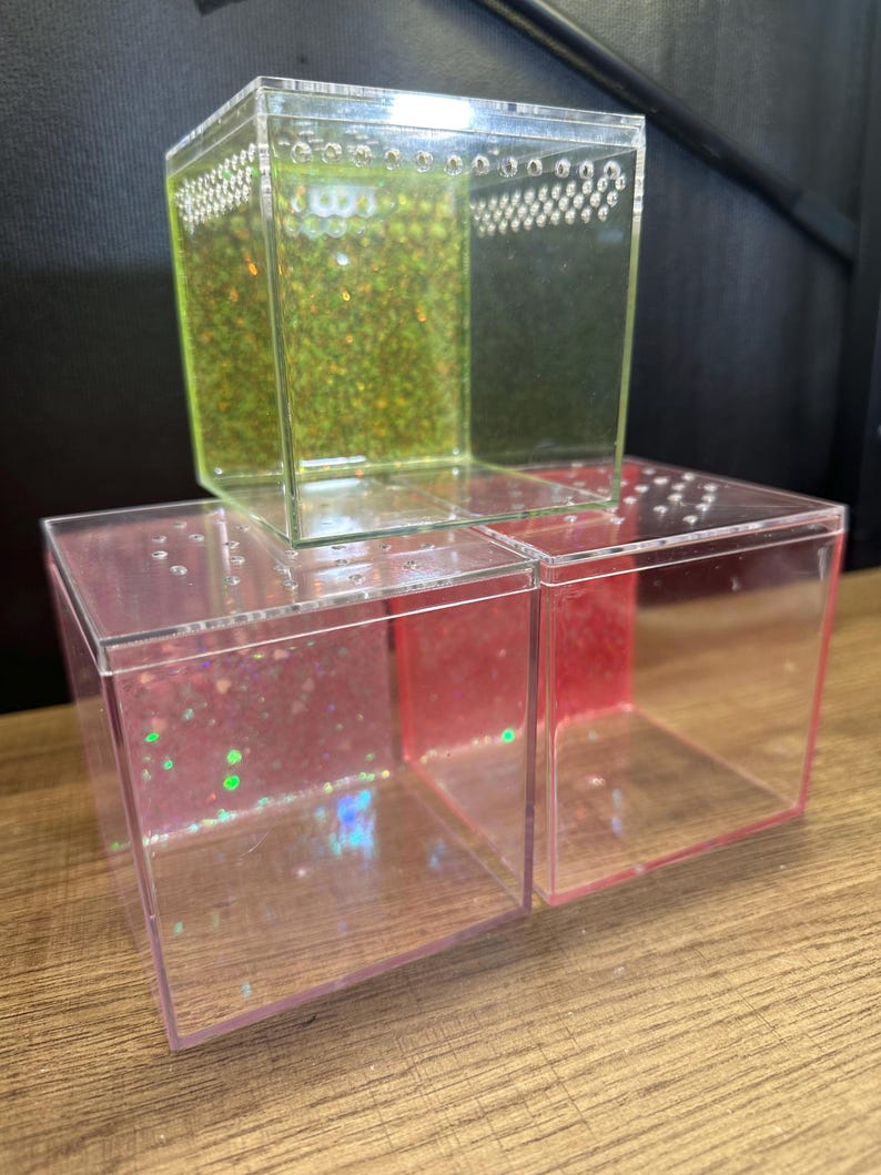 May include: Clear acrylic cube-shaped containers, one stacked on top of two others. The top cube has a green glitter filling, while the bottom cubes have pink and red glitter. Each cube has small holes along the top edge.