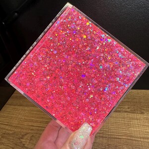 May include: A clear, square container filled with bright pink glitter. The glitter has an iridescent sheen, reflecting multiple colours. The container is held against a dark background and a wooden surface.