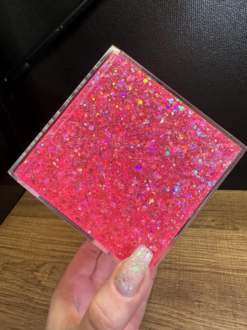 May include: A clear, square container filled with bright pink glitter. The glitter has an iridescent sheen, reflecting multiple colours. The container is held against a dark background and a wooden surface.