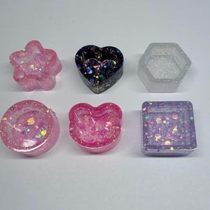 May include: Six small resin trinket dishes in various shapes including a star, heart, hexagon, circle, bear head, and square. Each dish features a unique finish with embedded glitter, iridescent flakes, and translucent or opaque color pigments.