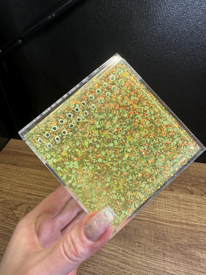 May include: A clear, square container filled with green and orange glitter. The top has a series of small, evenly spaced holes. The container is held in a hand, with a wooden surface in the background.