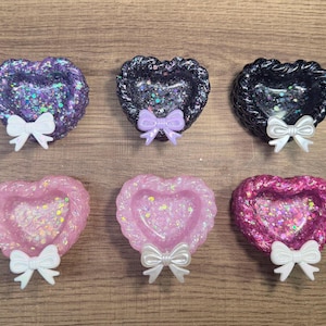 May include: Six heart-shaped trinket dishes in various colors, including purple, black, pink, and magenta. Each dish is filled with glitter and has a decorative bow attached. The dishes are arranged on a wooden surface.