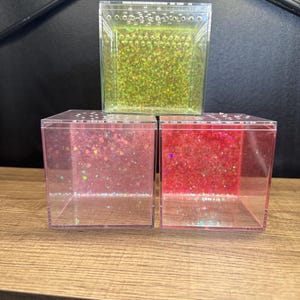 May include: Three clear, square containers filled with glitter. The top container has green glitter, while the bottom two have pink and red glitter. The containers are stacked on a wooden surface.