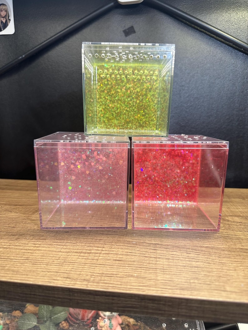 May include: Three clear, square containers filled with glitter. The top container has green glitter, while the bottom two have pink and red glitter. The containers are stacked on a wooden surface.
