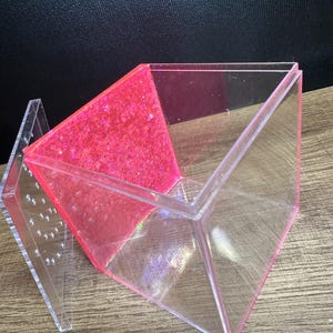 May include: A clear, geometric box with a hinged lid. One side is filled with bright pink glitter. The box is made of transparent acrylic and sits on a wooden surface. The lid is open, revealing the interior.