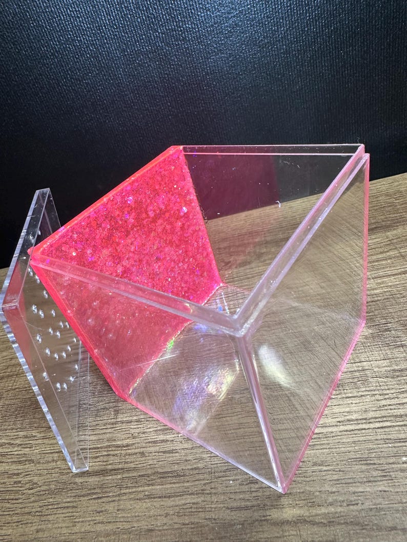 May include: A clear, geometric box with a hinged lid. One side is filled with bright pink glitter. The box is made of transparent acrylic and sits on a wooden surface. The lid is open, revealing the interior.