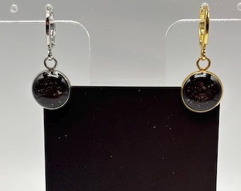 Puede incluir: Un par de pendientes redondos con un interior oscuro y moteado. Un pendiente tiene un aro plateado y el otro un aro dorado. Los pendientes se exhiben sobre una superficie negra con la palabra "MIDNIGHT" visible.