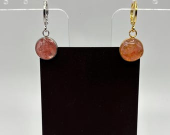 Puede incluir: Un par de pendientes con piedras redondas de color rosa rojizo. Uno tiene un aro plateado y el otro un aro dorado. Se exhiben en una tarjeta negra con el logotipo "Midnight Timber Co.".