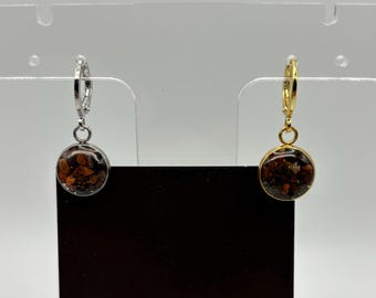 Puede incluir: Un par de pendientes con un colgante redondo de resina transparente lleno de pequeñas piedras de color marrón oscuro. Un pendiente tiene un aro plateado y el otro un aro dorado. Los pendientes se exhiben en un soporte transparente.