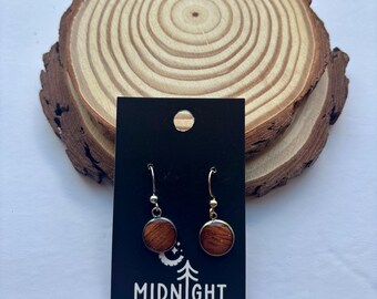 Puede incluir: Pendientes redondos de madera con ganchos plateados y detalles dorados, exhibidos en una tarjeta negra con "MIDNIGHT TIMBER CO." y un diseño de árbol. El fondo muestra una rodaja de madera con vetas naturales.