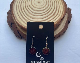 Puede incluir: Un par de pendientes colgantes plateados con incrustaciones redondas de madera oscura. Los pendientes se exhiben en una tarjeta negra con el texto "MIDNIGHT TIMBER CO." y un logotipo de luna creciente y árbol. El fondo es una rodaja de madera.