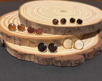 Puede incluir: Colección de pendientes de madera en varios colores y vetas, incluyendo marrón claro, marrón oscuro y rojizo. Los pendientes se exhiben sobre rodajas de madera rústicas, mostrando sus texturas naturales y engastes metálicos dorados.