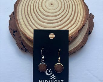 Puede incluir: Un par de pendientes redondos de madera con borde dorado y ganchos plateados, exhibidos en una tarjeta negra con el texto "MIDNIGHT TIMBER CO.". Los pendientes se presentan sobre un fondo de rodaja de madera natural.