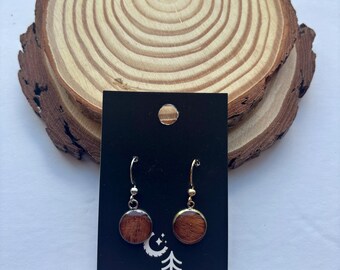 Puede incluir: Pendientes redondos de madera con ganchos plateados y dorados, presentados en una tarjeta negra con el logo "MIDNIGHT TIMBER CO.". Los pendientes se exhiben sobre un fondo de rodaja de madera natural. Diámetro: aprox. 1,5 cm.