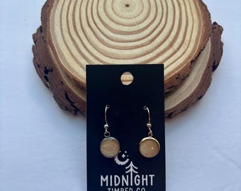 Puede incluir: Pendientes dorados con un diseño circular color arena, presentados en una tarjeta negra con el texto "MIDNIGHT TIMBER CO." Los pendientes se exhiben sobre una rodaja de madera natural con anillos de crecimiento visibles.