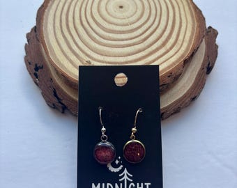Puede incluir: Un par de pendientes redondos de color rojo oscuro con ribetes y ganchos dorados, expuestos en una tarjeta negra con el texto "MIDNIGHT TIMBER CO." Los pendientes se presentan sobre un fondo de rodaja de madera natural.