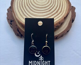 Puede incluir: Pendientes colgantes con colgantes circulares oscuros, presentados en una tarjeta negra con el texto "MIDNIGHT TIMBER CO.". Los pendientes se colocan sobre un fondo de rodaja de madera, creando un aspecto rústico. Los pendientes tienen ganchos plateados y dorados.