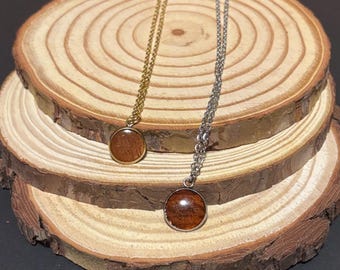 Puede incluir: Dos collares con colgantes redondos de madera. Uno tiene una cadena dorada y el otro una cadena plateada. Los colgantes son de color marrón rojizo y están engastados en marcos metálicos. Los collares se exhiben sobre una rodaja de madera.