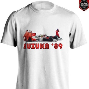 May include: White t-shirt with a racing car graphic and the text "SUZUKA '89" in red. The design shows a race car with the "Marlboro" logo, pit crew, and a driver in red. The shirt has a crew neck and short sleeves, suitable for casual wear.