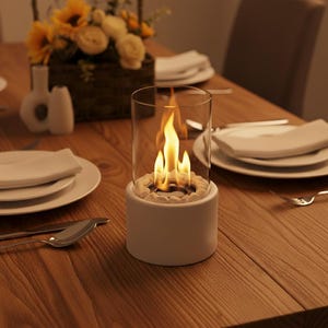 Peut inclure: Un foyer de table blanc avec un cylindre en verre et une flamme à l'intérieur. Le foyer est posé sur une table en bois, avec des assiettes, des couverts et une composition florale en arrière-plan.