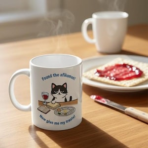 Może przedstawiać: Biały ceramiczny kubek z rysunkiem kota z kreskówki i napisem "Found the afikomen! Now give me my treats!". Na zdjęciu widoczny jest również talerz z krakersami z dżemem i biały kubek.