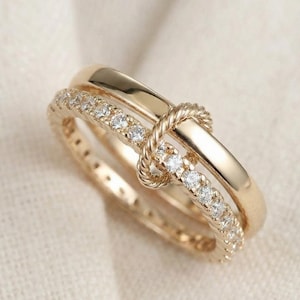 May include: A gold-colored ring set featuring two rings. One ring is a solid band, and the other is adorned with small, clear gemstones. The rings are intertwined with a rope-like design, creating a unique knot detail.