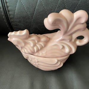 May include: A light pink ceramic rooster-shaped trinket box. The lid features a stylized crest, and the body is detailed with feather-like patterns. The box is designed with a handle on the side and is likely used for decorative storage.
