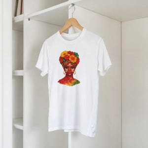 Puede incluir: Camiseta blanca con una vibrante ilustración de una persona con un turbante floral y pendientes de aro dorados. La obra de arte presenta un diseño colorido con tonos rojos, amarillos y verdes, creando un efecto visual llamativo en la camiseta.