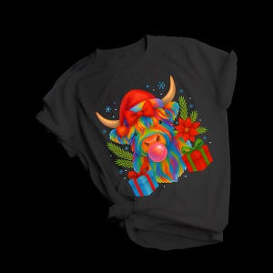 Puede incluir: Camiseta negra con una colorida ilustración de una vaca de las Tierras Altas con un gorro de Papá Noel y soplando una burbuja de chicle. El diseño incluye regalos de Navidad y vegetación festiva, ideal para la temporada navideña.