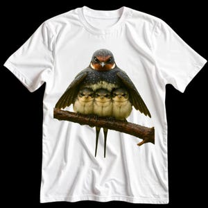 Puede incluir: Camiseta blanca con una imagen detallada de una golondrina protegiendo a tres polluelos en una rama. El ave adulta tiene plumas de color marrón oscuro y blanco moteado, mientras que los polluelos tienen plumas de color marrón claro y blanco.