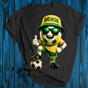 Puede incluir: Camiseta negra con un personaje de balón de fútbol de dibujos animados que lleva gafas de sol y un sombrero con la palabra "BRASIL". El personaje hace una señal de aprobación y está de pie sobre un balón de fútbol. El personaje lleva un uniforme amarillo y verde.