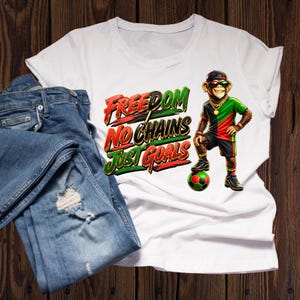 Puede incluir: Camiseta blanca con el texto "FREEDOM NO CHAINS JUST GOALS" en rojo, verde y amarillo. La camiseta presenta un mono de dibujos animados con gafas de sol, gorra y cadena de oro, junto a un balón de fútbol. Vaqueros azules doblados junto a la camiseta.