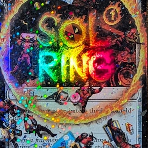 May include: A holographic trading card featuring Deadpool characters and the words "Soul Ring" in a rainbow gradient. The card has a black background with colorful accents and illustrations of Deadpool in various action poses.