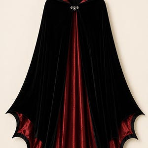 May include: A floor-length hooded cloak featuring a black velvet exterior and a deep red satin lining. The garment has a scalloped hemline and is secured at the neck with a silver-toned metal clasp featuring a central circular accent piece.