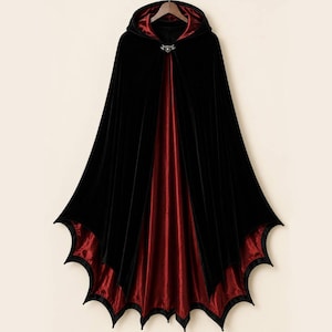 May include: A floor-length hooded cloak featuring a black velvet exterior and a deep red satin lining. The hem is cut into a distinct scalloped bat-wing shape. A silver-toned metal clasp with a central circular detail secures the garment at the neck.