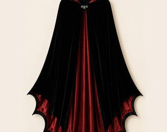 May include: A floor-length hooded cloak featuring a black velvet exterior and a deep red satin lining. The hem is cut into a distinct scalloped bat-wing shape. A silver-toned metal clasp with a central circular detail secures the garment at the neck.