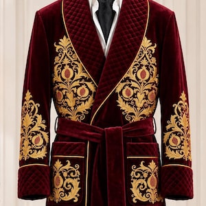 May include: A floor-length burgundy velvet smoking robe featuring gold embroidered pomegranate motifs on the chest and sleeves. The robe includes a quilted shawl collar, matching tie belt with gold tassels, and two front pockets with gold piping.
