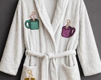 May include: White plush robe with embroidered coffee cup designs in green, purple, yellow, and red. The robe has a shawl collar, a waist tie, and two front pockets. The robe is hanging on a black hanger.