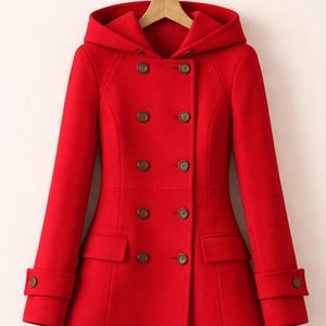 May include: A vibrant red double-breasted coat featuring a hood, long sleeves with buttoned cuffs, and two rows of dark brown buttons down the front. The coat includes two flap pockets at the waist and is displayed on a wooden hanger.