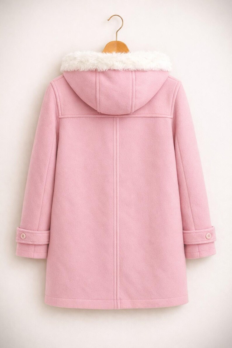 May include: A light pink duffel coat with a hood and white faux fur trim. The coat has a button closure and two buttons on the sleeves. The coat is hanging on a wooden hanger.