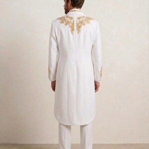 May include: A long white coat featuring intricate gold embroidery across the upper back and shoulders, paired with matching white trousers and white shoes. The coat has a structured collar, long sleeves with gold detailing at the cuffs, and a hem.